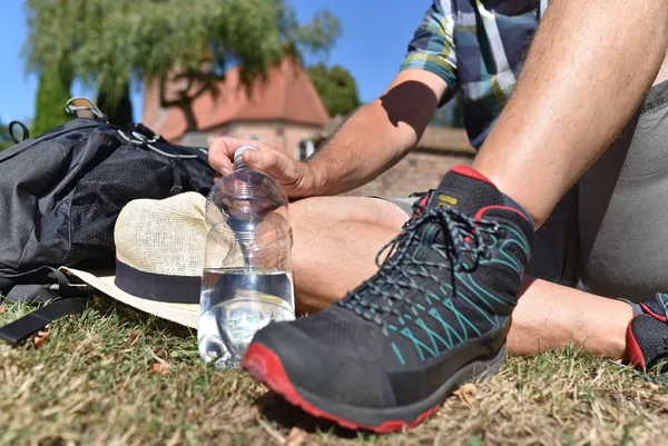 Ein Pilger macht eine Trinkpause; der Fokus liegt auf der Wasserflasche. Im Hintergrund ist verschwommen eine hübsche Kapelle mit Friedhof zu sehen.