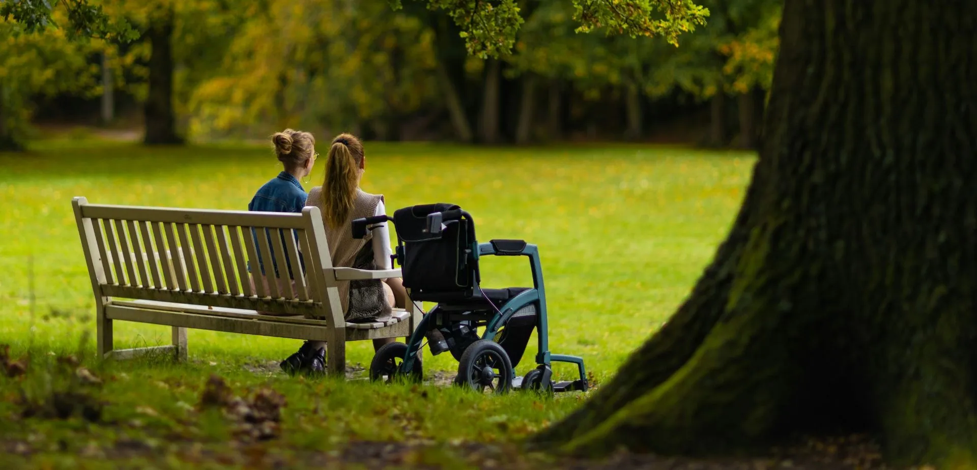 Zwei Frauen sitzen auf einer Parkbank, neben ihnen ein Rollstuhl