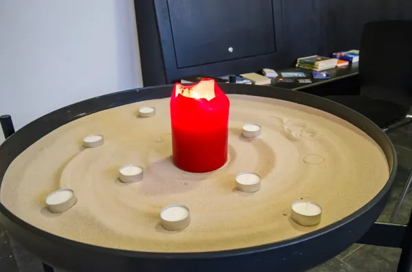 große Schale mit Sand. In der Mitte eine rote Kerze und außenherum Teelichter