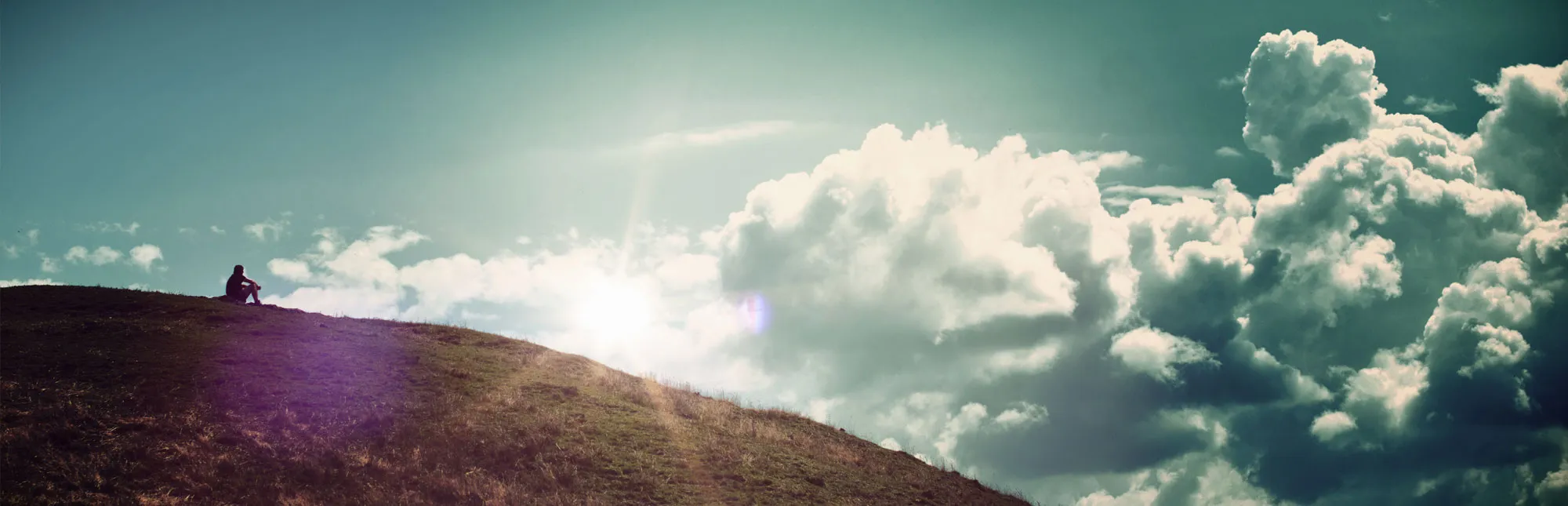Person sitzt auf einem Hügel und schaut in die Wolken
