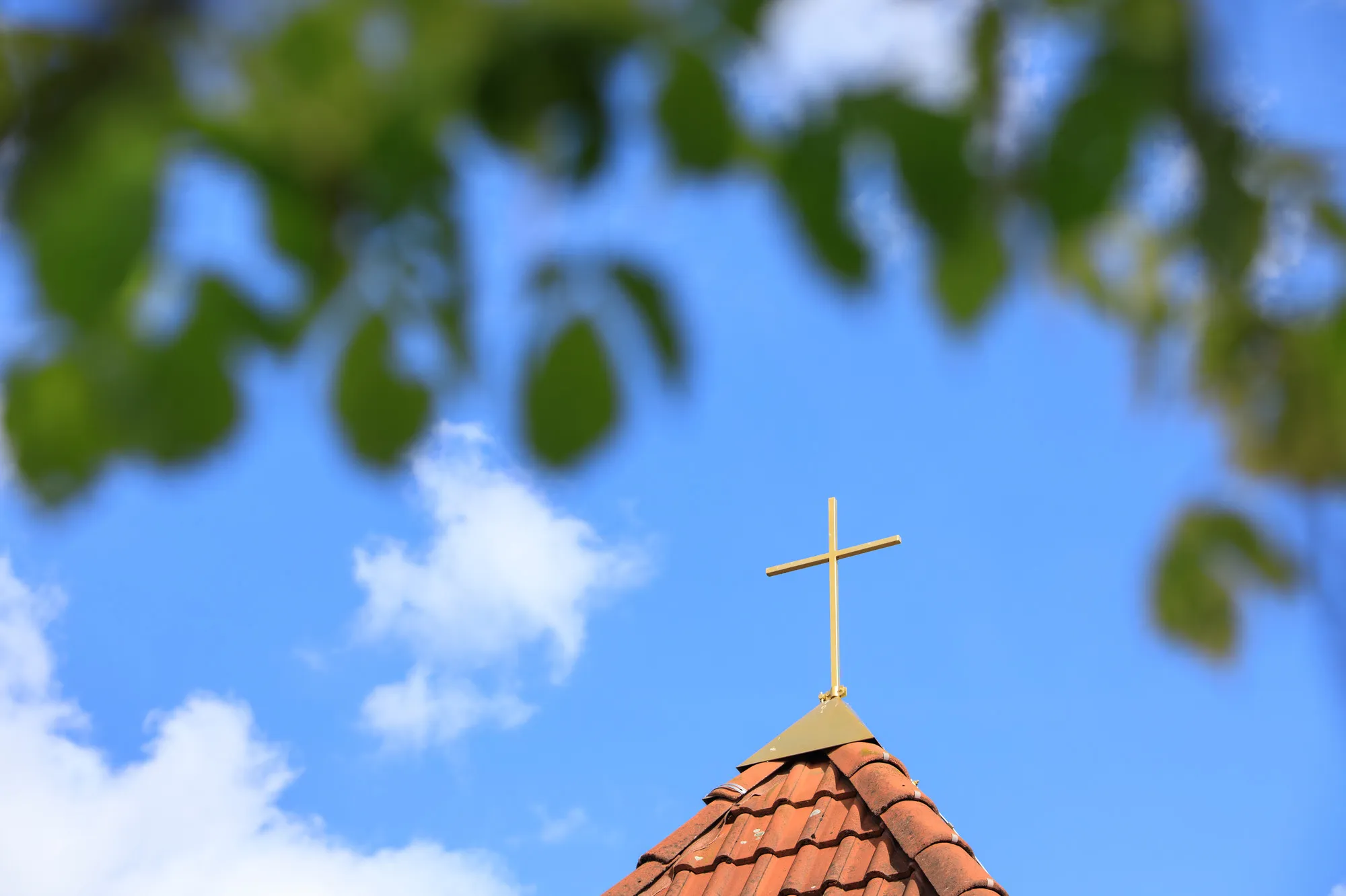 Kirchturmkreuz vor blauem Himmel, gerahmt von unscharfen Blättern eines Baums