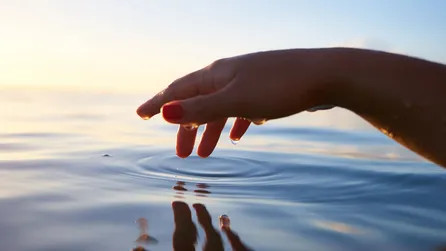 Hand kommt von rechtsin bild, darunter Wasser , die Hand nach unten gebogen und berührtdas Wasser, das an der Stelle einen leichten Kreis entstehen lässt.