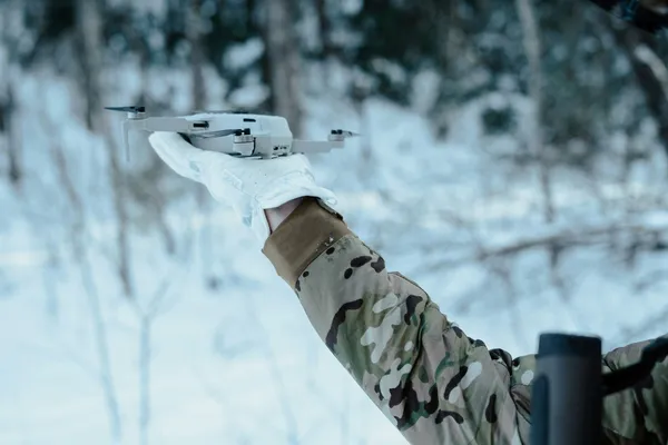 Winterlandschaft, eine Person in Flecktarnuniform hält eine kleine Drone in der Hand