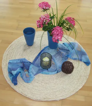 Bodenbild mit runder heller Bastmatte blaues Tuch wie Wasser darauf Kerze, Becher und Blumen