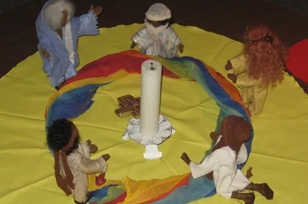 Rundes gelbes Tuch auf dunklem Untergrund. Darauf in der mitte eine Kerze und kleines Kreuz. Regenbogenfarbiges Tuch als Ring und Eglifiguren in unterschiedlichen Gebetshaltungen