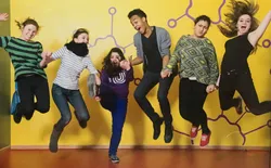 Jugendliche stehen auf eine Bühne, gelber Hintergrund , halten sich an den Händen und machen gemeinsam einen Luftsprung