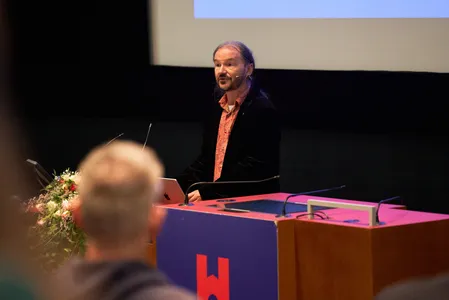 Gernot Meier hält einen Vortrag. Ein bärtiger Mann mit langen dunkelgrauen Haaren trägt einen dunklen Anzug