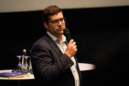 Tobias Kreilos hält einen Vortrag. Ein Mann mit kurzem braunem Haar trägt einen dunklen Anzug