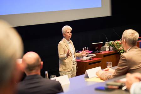 Publikum im Vordergrund hört die Rede von Marie-Agnes Strack-Zimmermann. Eine Frau mit kurzem weißen Haar trägt einen sandfarbenen Anzug