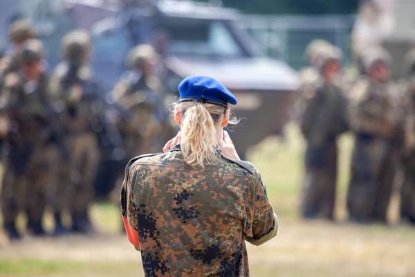 Eine Soldatin in Rückansicht, mit blonden Haaren, Pferdeschwanz, blauer Kappe und Tarnkleidung steht auf einem Übungsplatz. Im Hintergrund sind unscharf weitere Soldat:innen zu erkennen.
