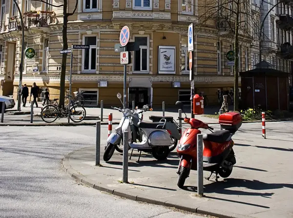 Straße macht eine Biegung nach Rechtsim Hintergrund ein großes gelb/weiß gestrichenes Haus. Im Vordergrund ein Platz abgetrennt von Str. auf dem 2 Motorroller geparkt sind