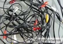 Auf hellgruem Hintergrund liegt ein Audio-Kabelsgwirr mit gelben, roten, scharzen und weißen Steckern die gleichzeitg etwas Schatten auf die hellgraue Fläche werfen. Darunter die Worte: " Gefühle entwirren"