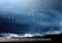Dunkler Wolkenhimmel mir großen Regentropfen, wie Glaskugeln. Am unteren Rand ein in schwarz gehaltener Bergkamm. Darunter die Worte: "Im Schweren Leichtigkeit entdecken"