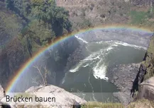 Blick von einer Felswand in die Tiefe auf einen Fluß. Auf der anderen Seite des Flusses ein Wald.So etwa auf 2/3 Höhe des Waldes ziegt sich ein Regenbogen im Halbkreis des Bildes. Darunter die Worte: " Brücken bauen.