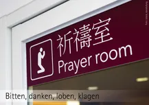 In einem Fenster hängt am oberen Rande queer über das Fenster ein rotes Schild. Links von eimem weiße rechteck ein Piktogramm vmit einem kieenden Männchen, die Arme zum Gebet erhoben, darüber in japanischer und englicher Schrift: Prayerroom. Darunter die Worte: "Bitten, danken, loben, klagen