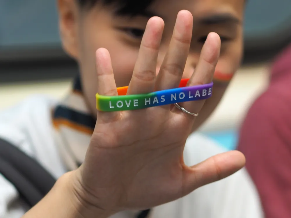 Person (unscharf zu sehen) zeigt ihre Hand mit einem Queer-Band, auf dem steht: "Love has no label"