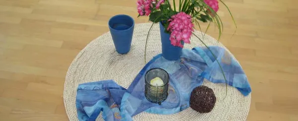 Spirituelle Mitte: Runde Unterlage aus Bast, ein blauen Tuch wie ein Fluß darüber gelegt, eine blaue Vase mit roten Blume drauf und Kerze im Glas und blauer Bechern auf Tischdecke